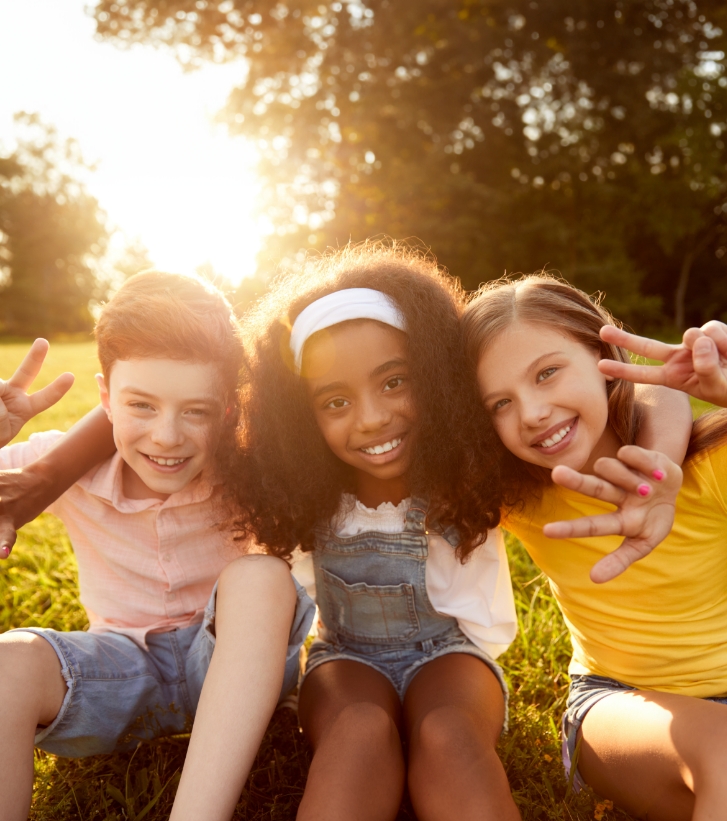 Three smiling kids sitting in grass and giving peace signs